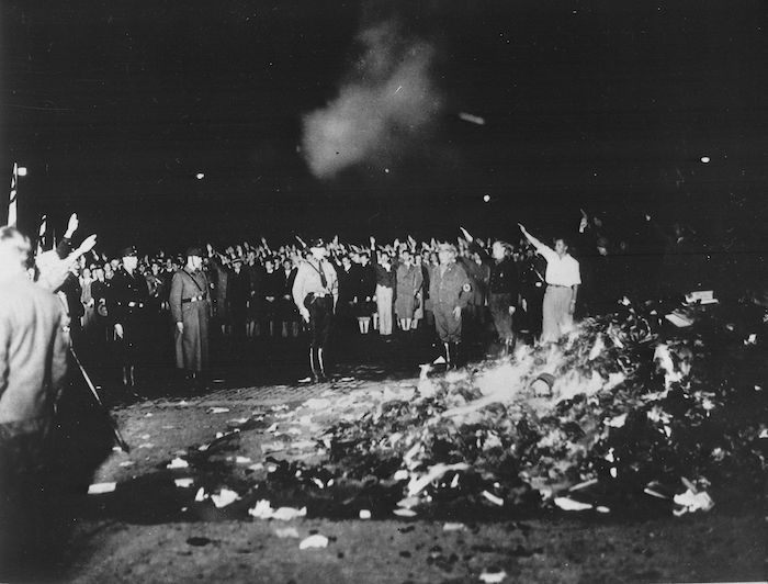 black and white image of a group of people performing the Nazi salute next to a large pile of burning photos