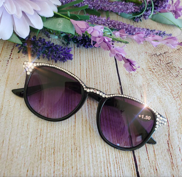 Black reading sunglasses with rhinestones along top next to purple flowers