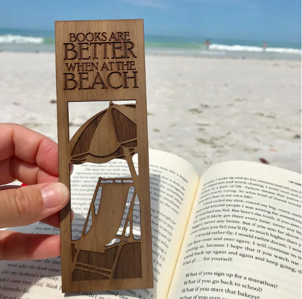 Wooden bookmark with umbrella, beach chair, and "Books are better at the beach" carved into it, held by a hand over a book with a beach background