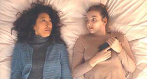 a still from killing Eve, showing Eve and Villanelle lying on their backs in a bed, looking at each other. Villanelle is holding a gun.