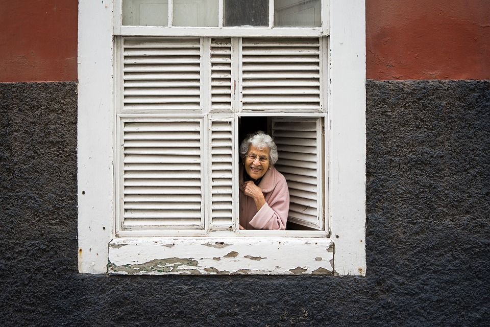 An older woman looks out of the bottom right corner of a window with white shutters. She has curly white hair, is wearing a pink robe, and is smiling.