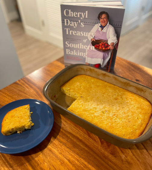 A rectangular pan full of cornbread sits on a wooden table in front of Cheryl Day's Southern Treasury of Baking. One square slice has been removed from the pan and is on a blue plate to the side of the pan.