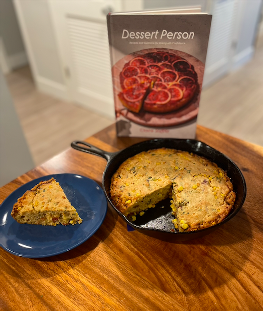 A large cast iron skillet of cornbread with visible bacon and peppers mixed in sits on a wooden table. One slice is set next to the pan on a blue plate, and behind the cornbread is Claire Saffitz' Dessert Person cookbook.