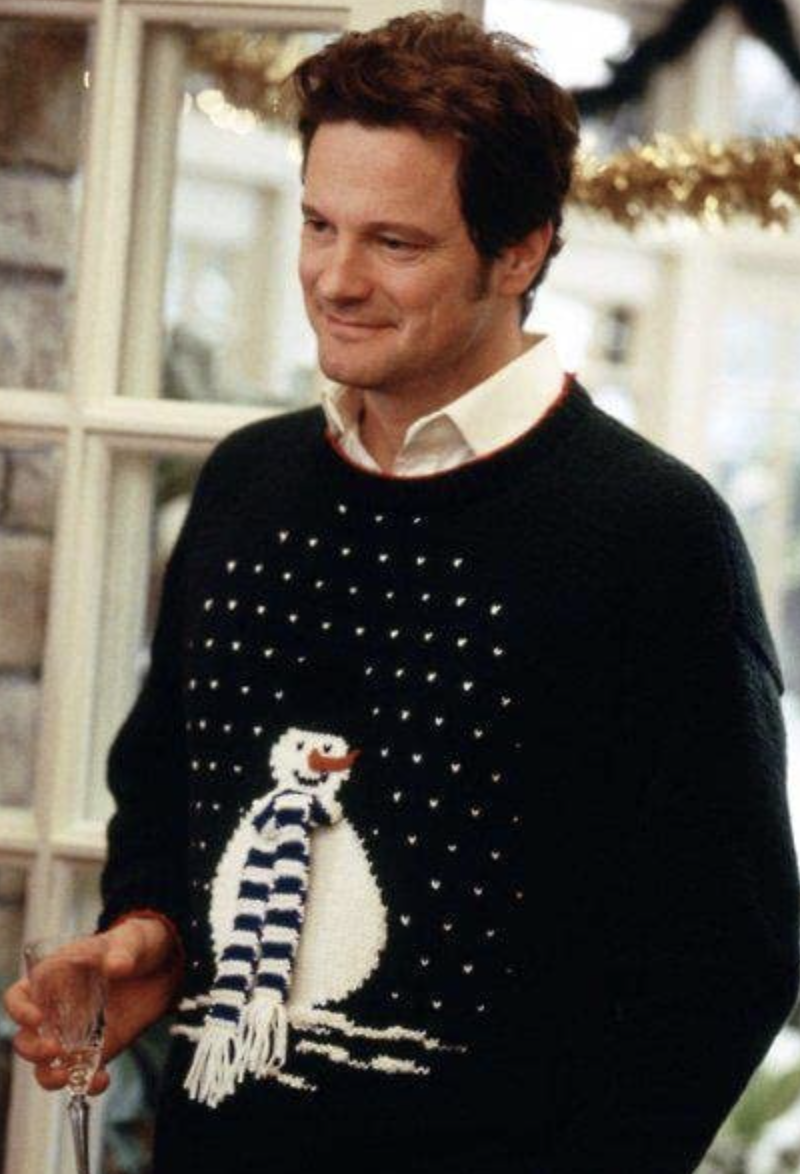 Colin Firth as Mark Darcy is wearing a sweater with a snowman on it and smiling while holding a glass of champagne.