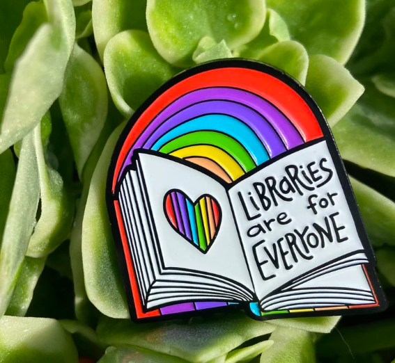 Enamel pin shaped like a book and rainbow that says "Libraries are for everyone". The pin is sitting on top of a succulent.