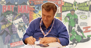 a photo of Neal Adams signing prints at a convention with his comic covers in the background