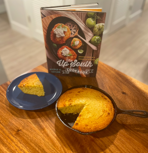 A cast iron skillet of golden brown cornbread on a wooden table. One slice is removed and next to the skillet on a blue plate, and behind the cornbread is Nicole A. Taylor's The Up South Cookbook.