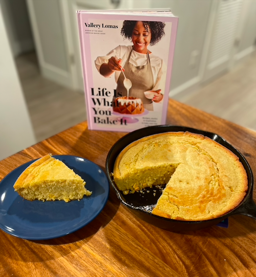 A large cast iron skillet of yellow cornbread is on a wooden table in front of Vallery Lomas' Life is What You Bake It cookbook. One slice of the cornbread has been set on a blue plate next to the skillet.