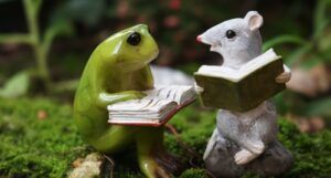 Two tiny statues of a green frog and a white mouse, both holding open books, sit on a bed of moss.