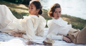 a photo of two women in old-fashioned clothing laying on a blanket at a park, with lilacs and a vintage book