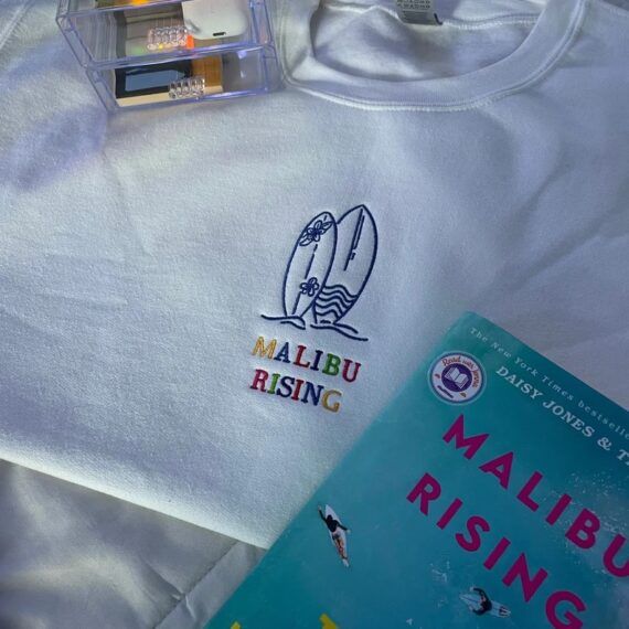 White crewneck with an embroidery of the words "Malibu Rising" and two surfboards