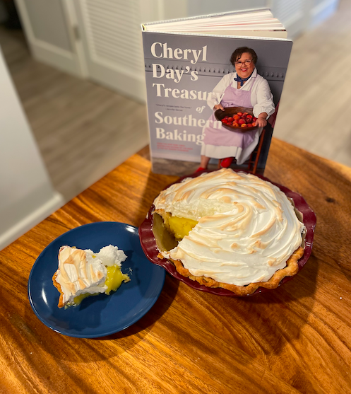 A lemon meringue pie sits on a wooden table with one slice, looking a little messy, sits on a blue plate to the side. Behind it is the cookbook Cheryl Day's Treasury of Southern Baking