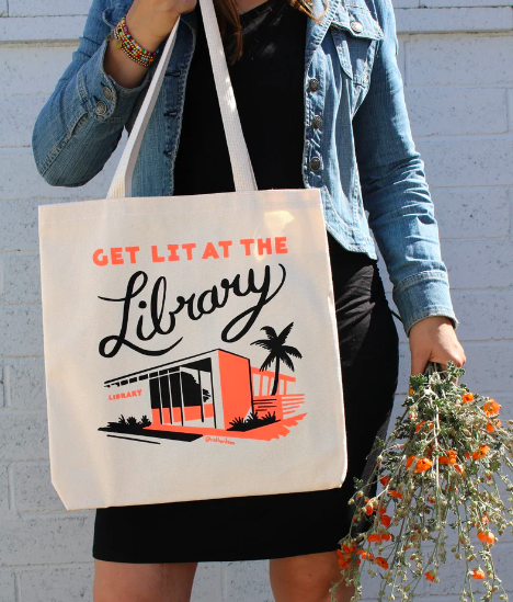 A person holding a beige tote bag with a retro graphic of a library that says "Get lit at the library"