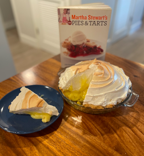 Photo of a lemon meringue pie on a wooden table. Once mess slice is on a blue plate. Behind the pie is the cookbook Martha Stewart's New Pies and Tarts.
