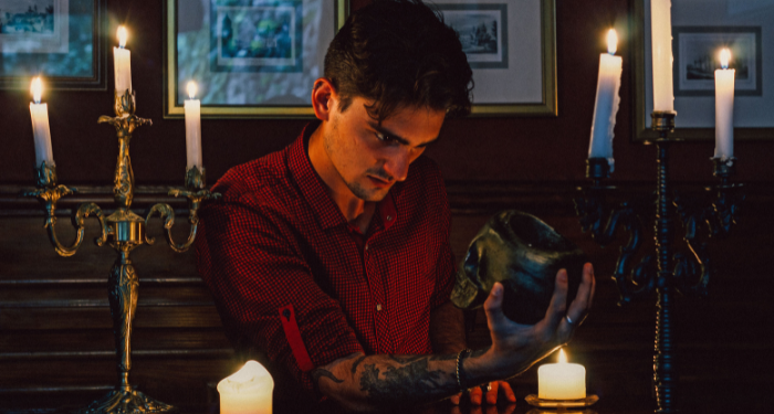 a young man with tattoos staring at a skull, surrounded by candles