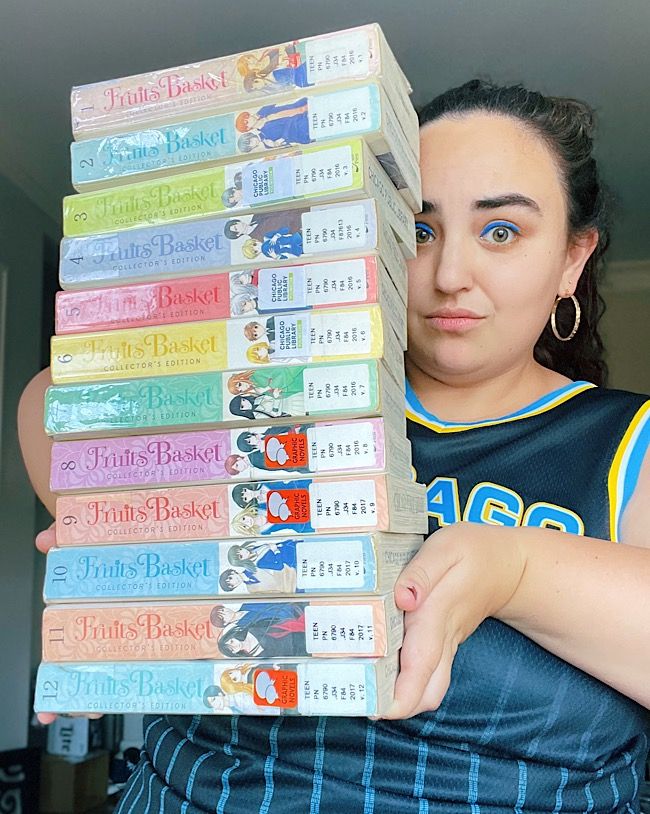 Leah, a woman with brown hair, wearing a basketball jersey, holding all 12 volumes of the Fruits Basket manga