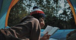 person reading outdoors in a tent. They are wearing a beanie and reading on their stomach
