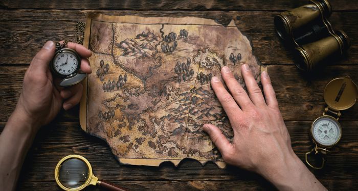 a pair of hands spreading out a treasure map with pieces missing. One hand is holding a pocket watch, off to the sides are a set of binoculars and a magnifying glass