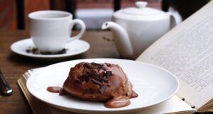 a chocolate dessert ona white plate on top of an open book with a white cup, saucer, and teapot in the background