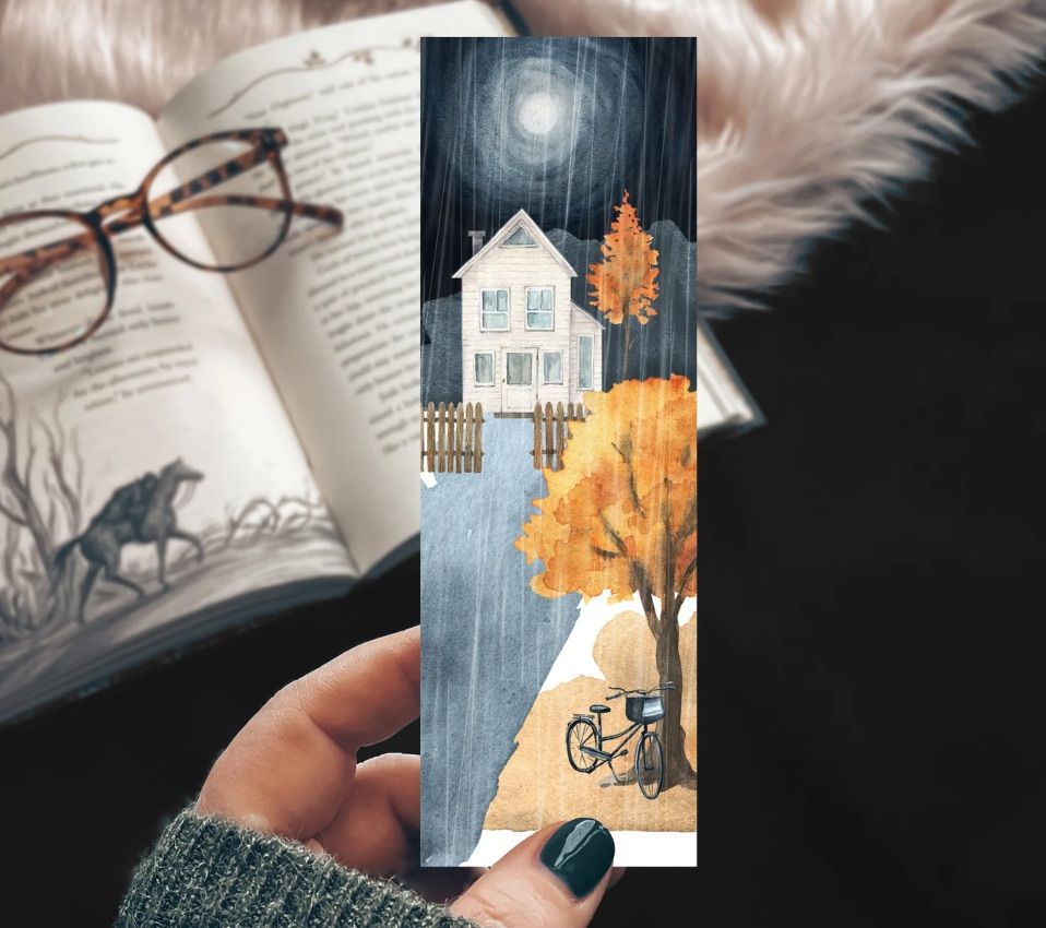 Image of an olive skin hand holding a bookmark. The bookmark features an older house, moonlight, rain, colorful fall trees, and a bike. 