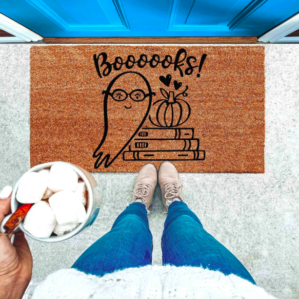 A brown doormat with a black outlined of a ghost in glasses next to a stack of books topped with a pumpkin and the text "Booooooks!"