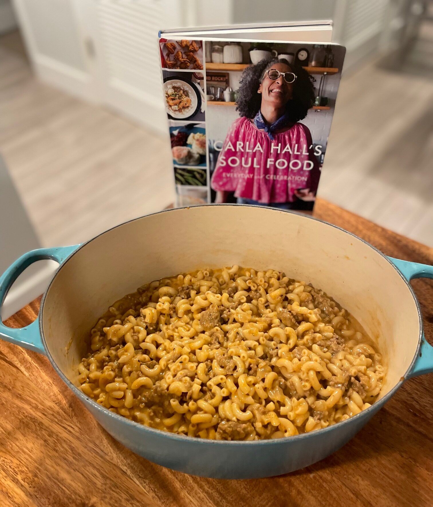 A teal dutch oven with a beefy mac and cheese inside, on a wooden table in front of the cookbook Carla Hall's Soul Food