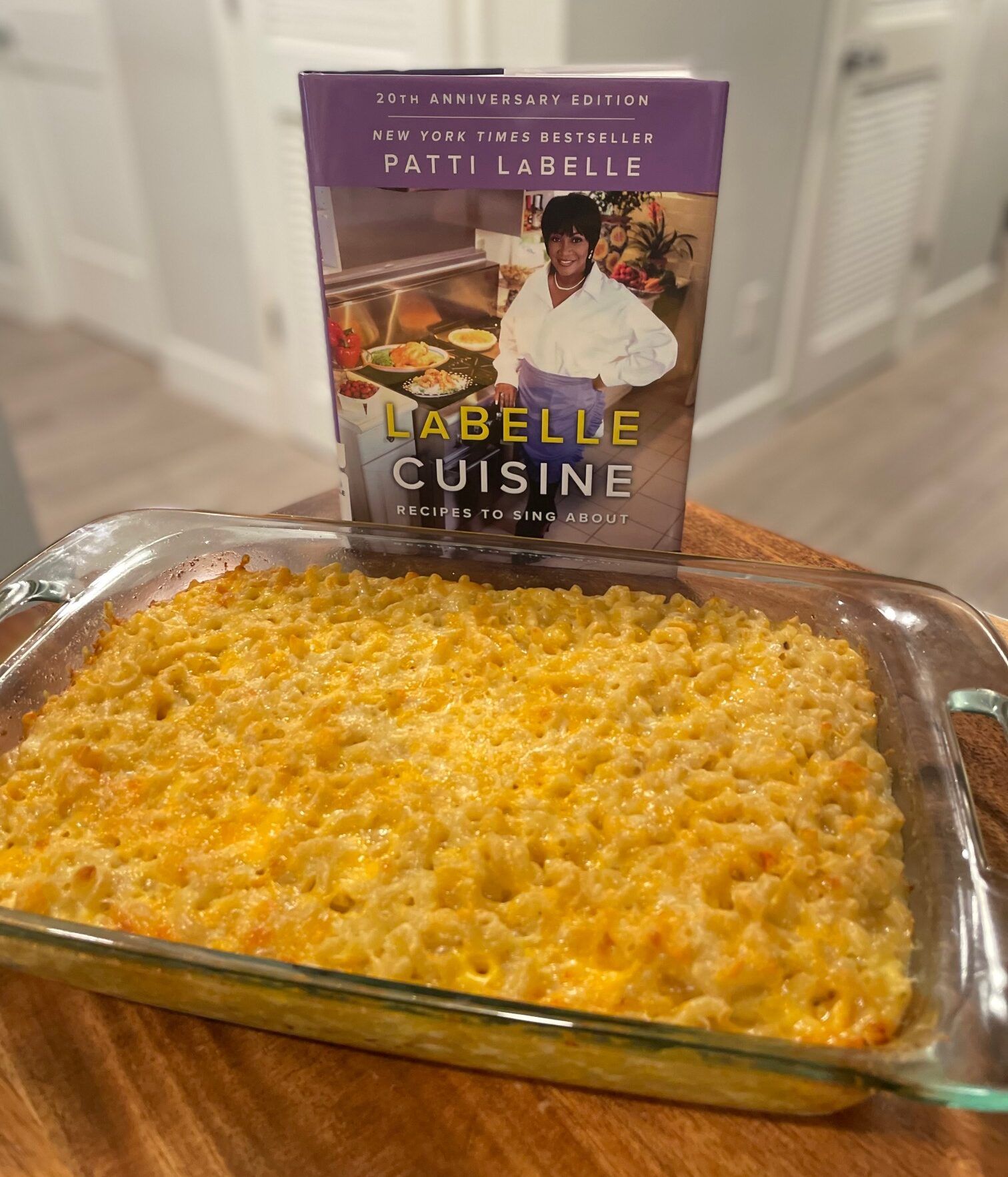 A glass casserole dish full of baked mac and cheese on a wooden table in front of the cookbook LaBelle Cuisine