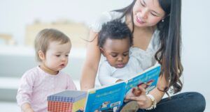 light-skinned Asian woman reads to a brown-skinned Black toddler, and a fair-skinned white baby