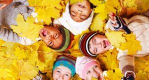 5 kids of differing skin tones lying in a pile of yellow leaves smiling