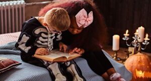 A little white boy in a skeleton costume and Black girl with a box in her hair are both reading the same book; there is a jack-o-lantern on the floor