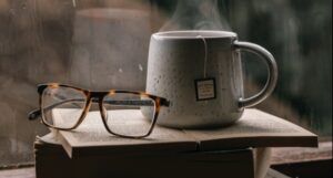 a steaming cup of tea perched on a stack pof books near a window next to a pair of tortoise shell glasses