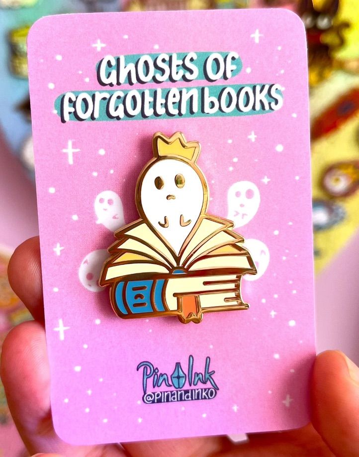 Image of an enamel pin with books and a ghost emerging from one of the open books.. 