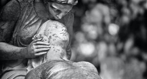 a black and white photo of a statue of someone kissing the other's forehead. The statues are beginning to detiorate.