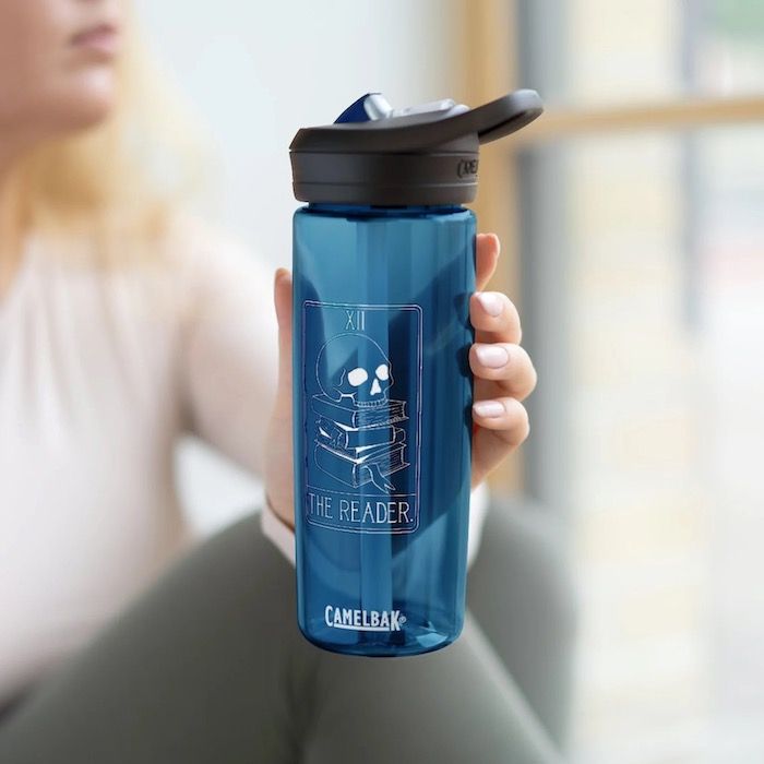 a blue CamelBak water bottle with a straw lid with a tarot card design. The card is a pile of books with a skull ob top and with text above it that says "The Reader"