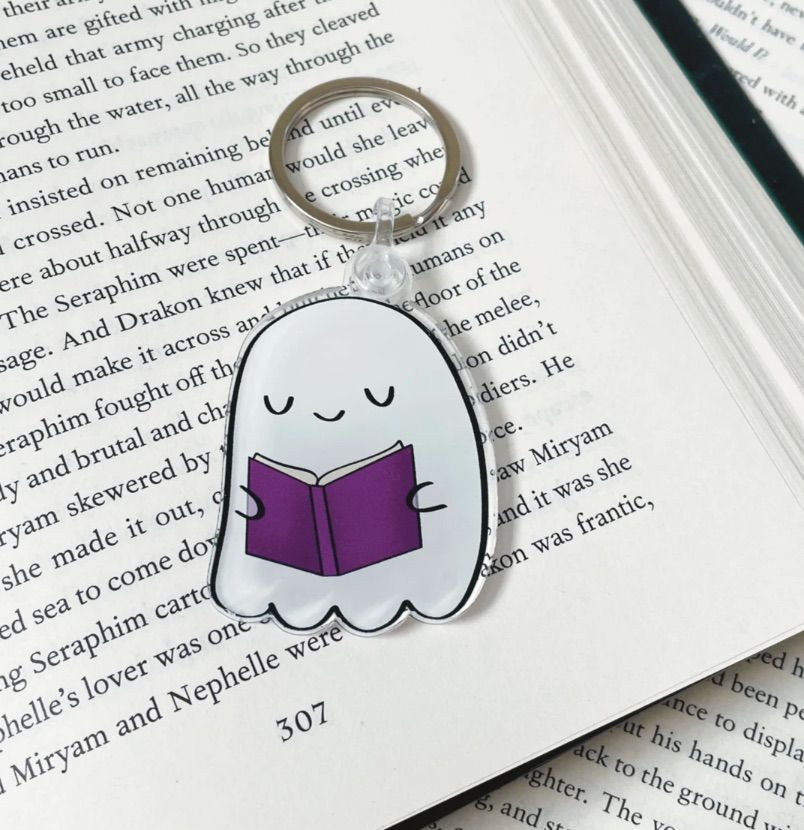 Image of a keychain in the shape of a ghost. It is reading a purple book. 