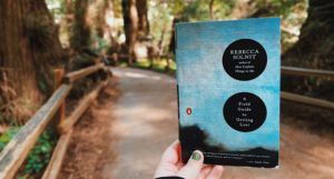 a fair-skinned hand holds the Rebecca Solnit’s A Field Guide to Getting Lost on a nature trail