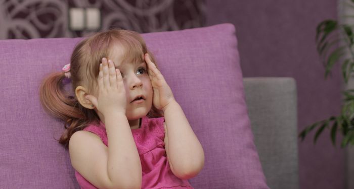 a small child sitting on a purple couch while watching TV, hands held up to their face in a scared expression