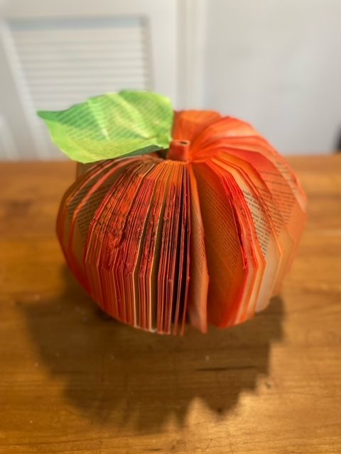 A 3D orange pumpkin made from an old book carved and fanned out. On top is a green leaf made from an old book page.
