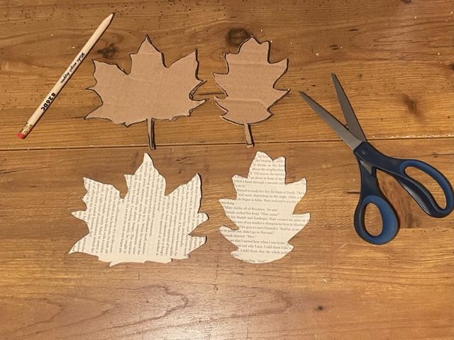Two cardboard leaf stencils next to leaf shapes cut from book pages on a wooden table next to scissors and a pencil