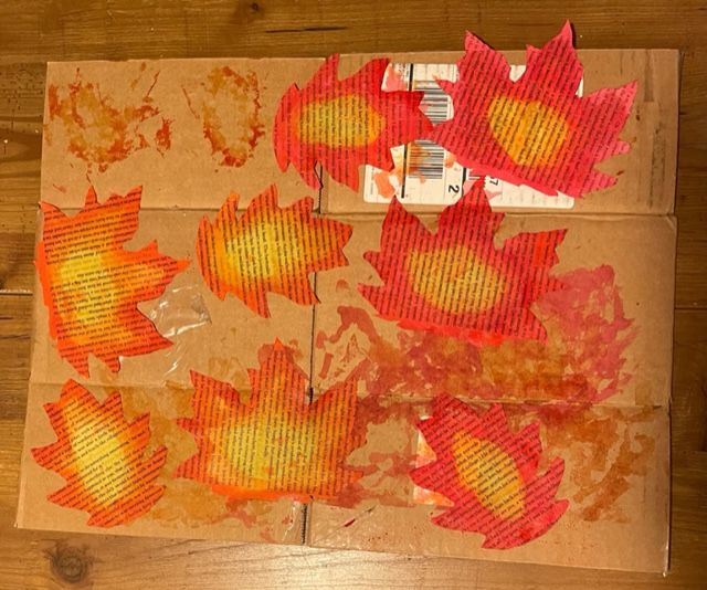 Eight colorfully painted leaves from old book pages are drying on a piece of cardboard