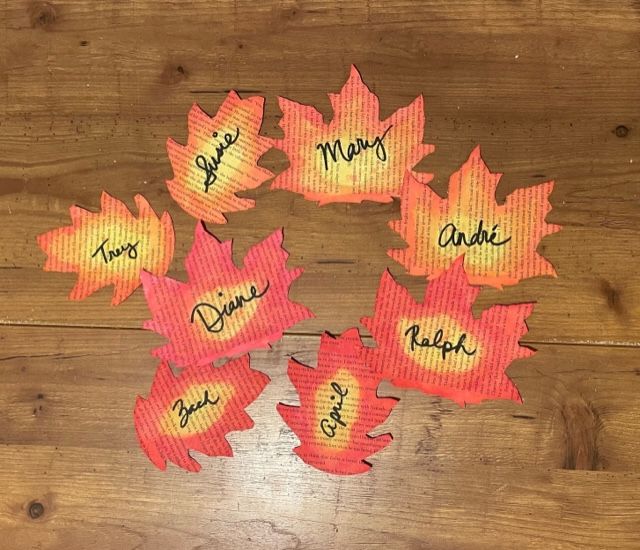 Eight colorfully painted leaves cut from book pages are on a wooden table. Names have been written with black marker in cursive in the center of the leaves.