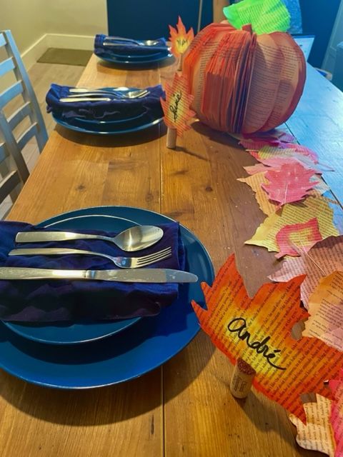 Photo of a set table with a colorful leaf garland made from book pages, a book page leaf with the name André in the foreground, blue plates and silverware, and a 3D book pumpkin made from a carved book