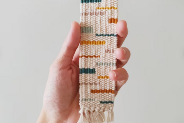 A hand holding up a cream colored hand-woven bookmark with autumnal colored horizontal dashes of varying lengths. 