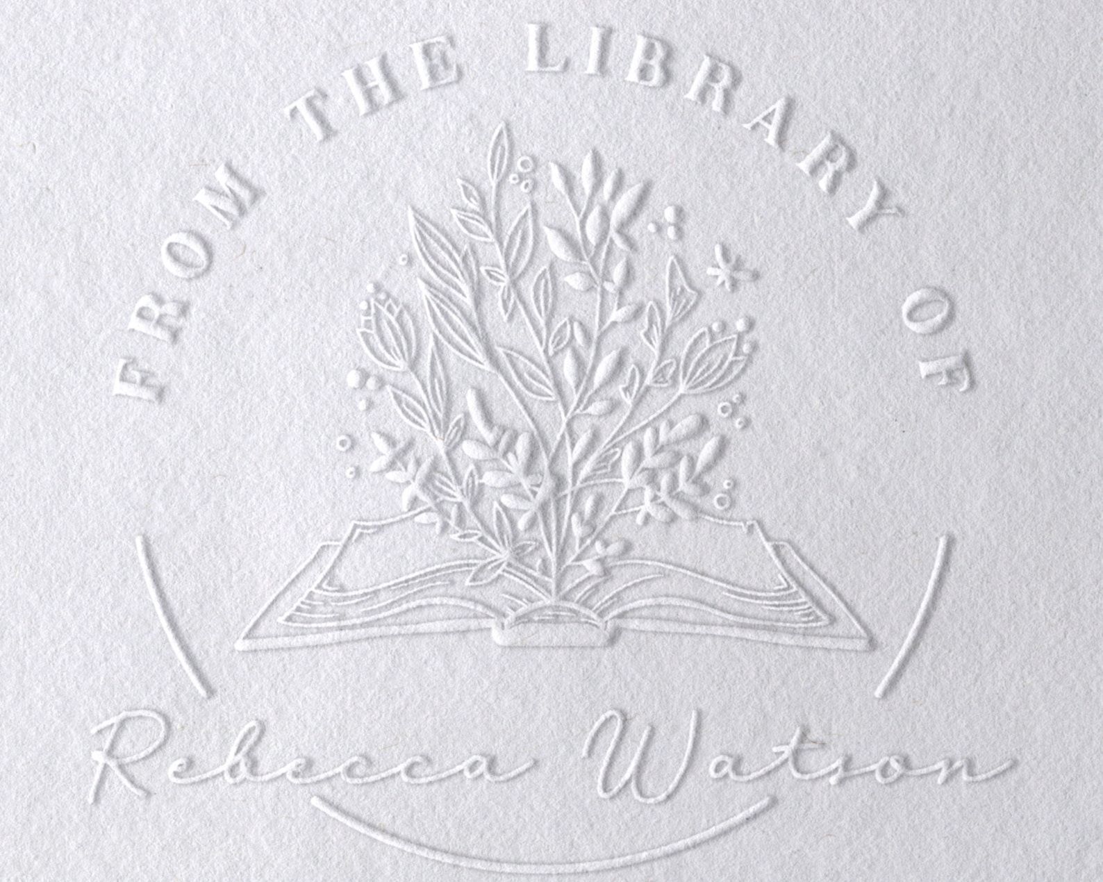 A white page wooing an embossed stamp that reads "from the library of ________" and an open book with flowers