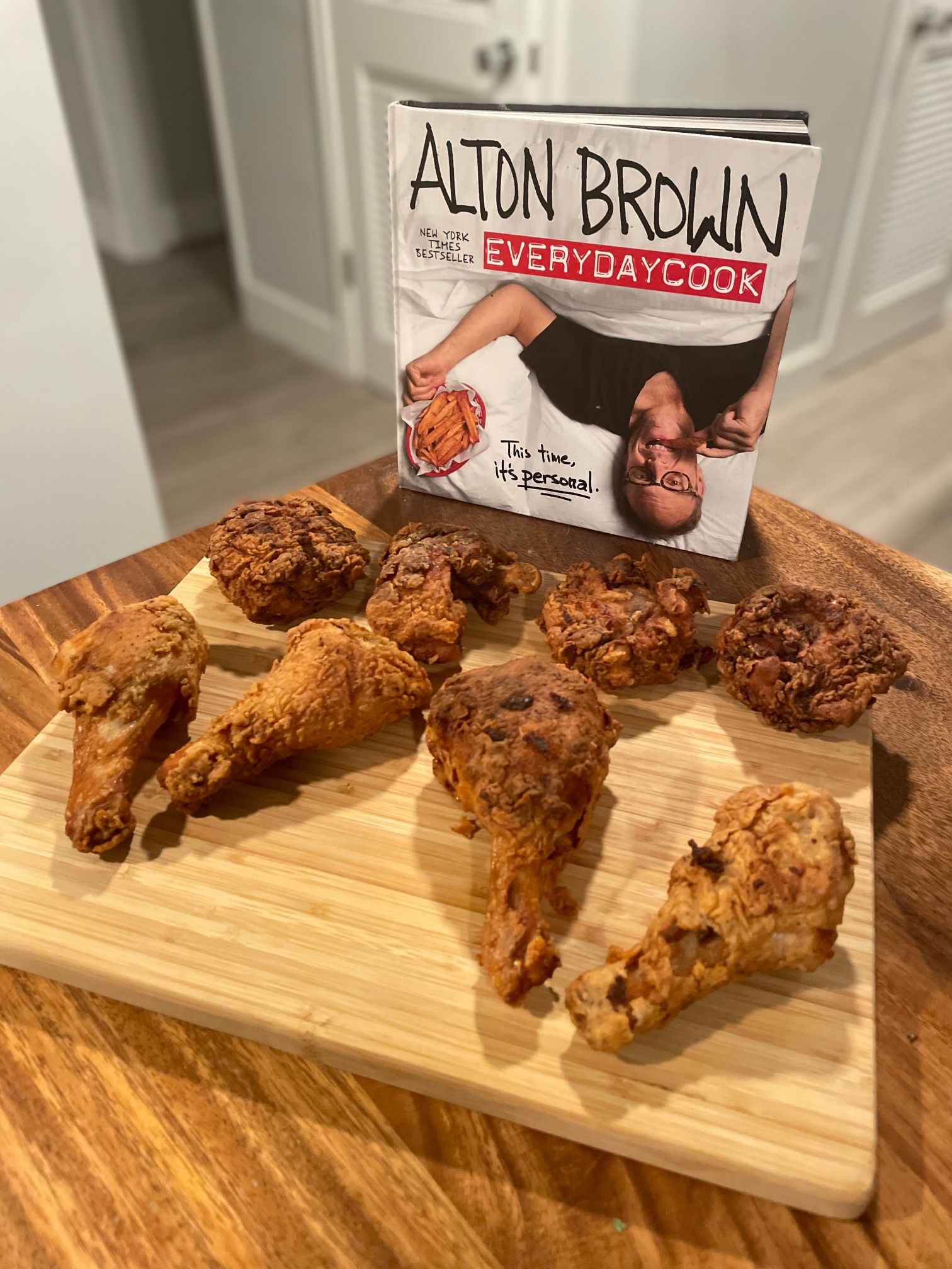 Fried chicken thighs and legs sit on a wooden cutting board in front of the cookbook EveryDayCook by Alton Brown
