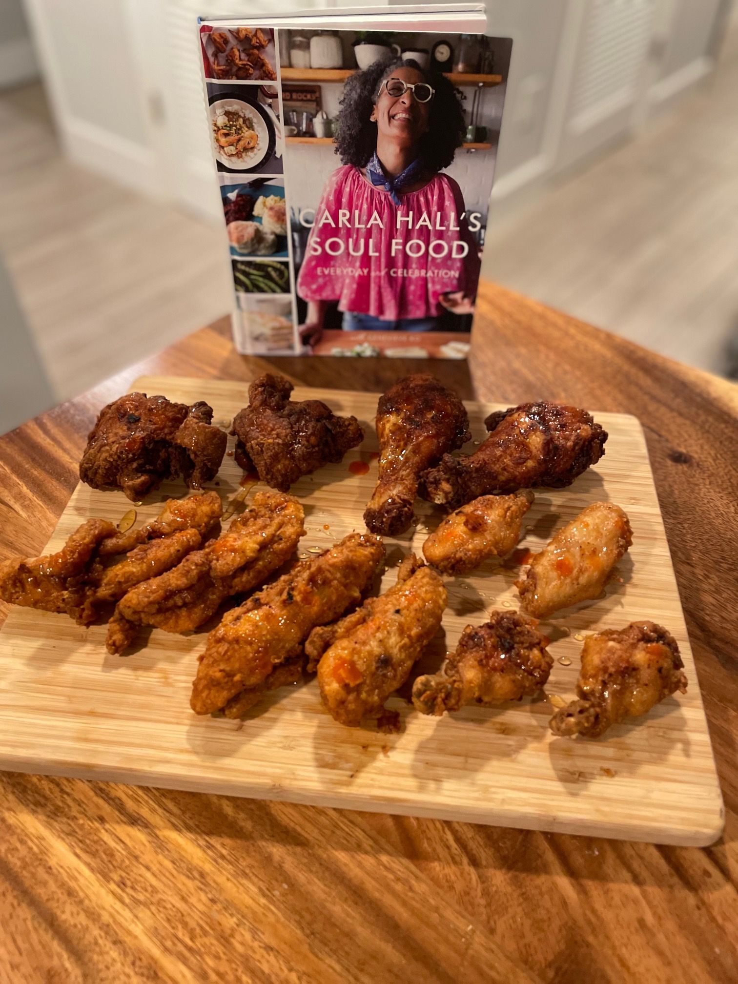 Various fried chicken pieces sit on a wooden cutting board in front of the cookbook Carla Hall's Soul Food