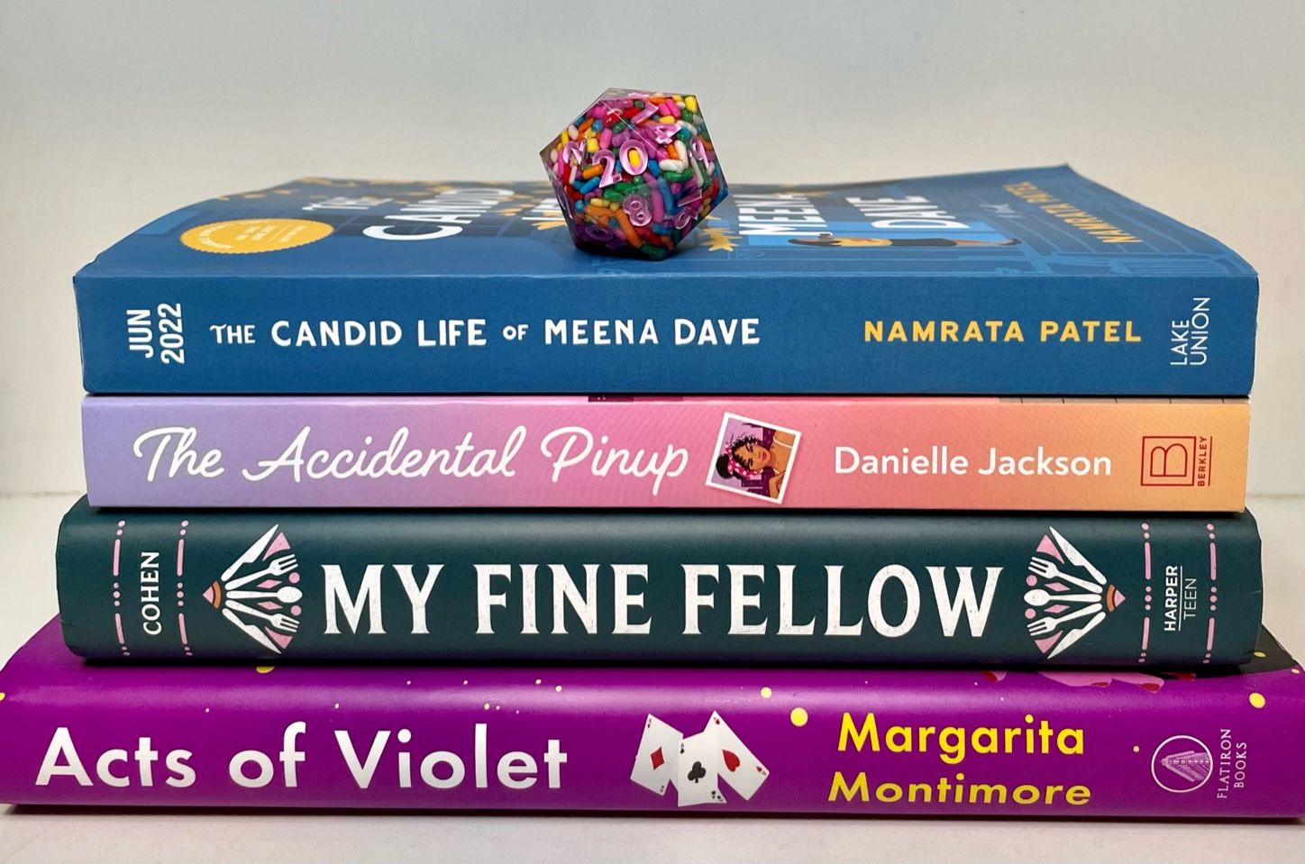 A 20-sided die full of sprinkles with pink numbers sits atop a stack of books (The Candid Life of Meena Dave, The Accidental Pinup, My Fine Fellow, and Acts of Violet)