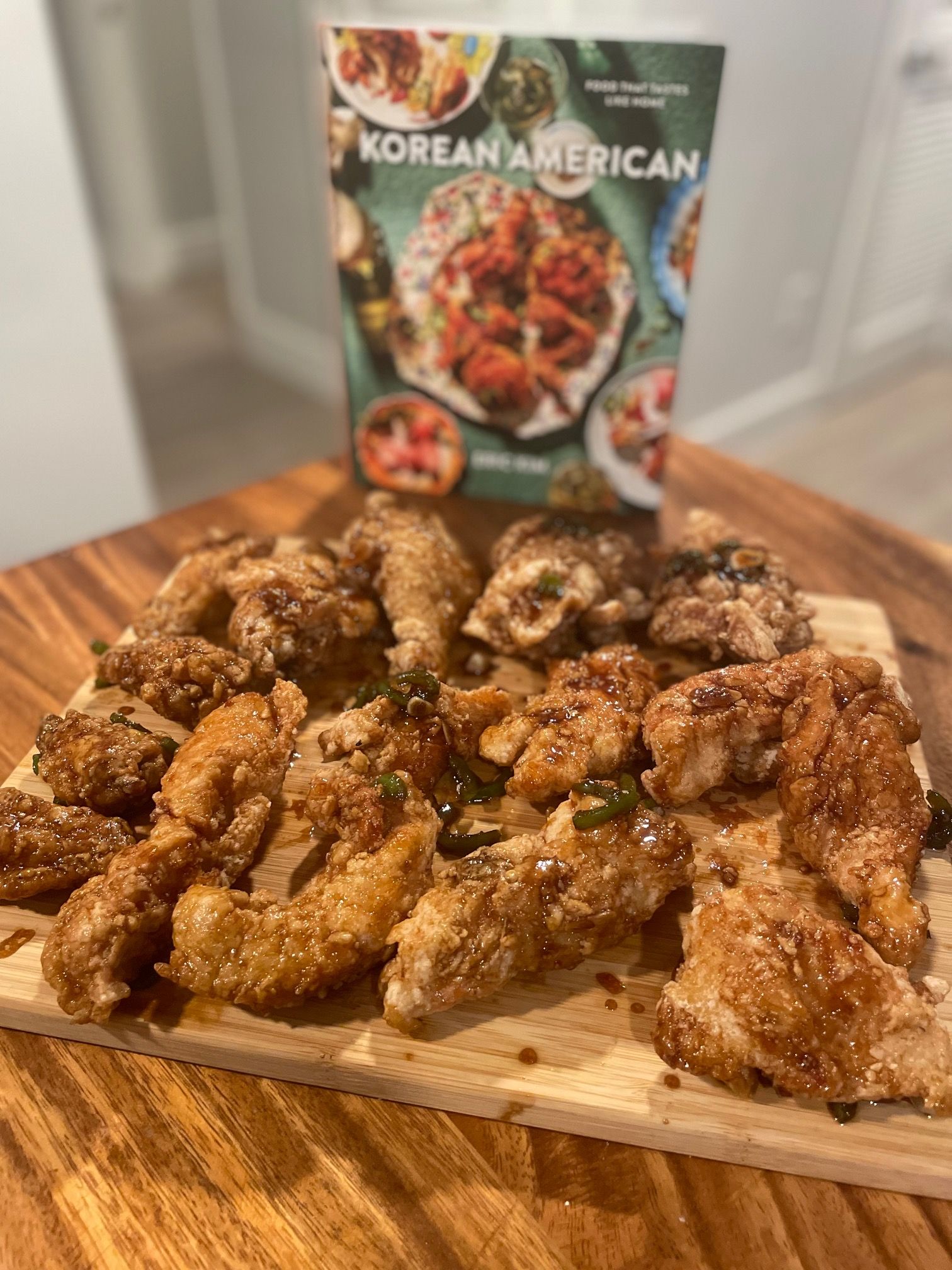 Various fried chicken pieces on a wooden cutting board in front of the cookbook Korean American