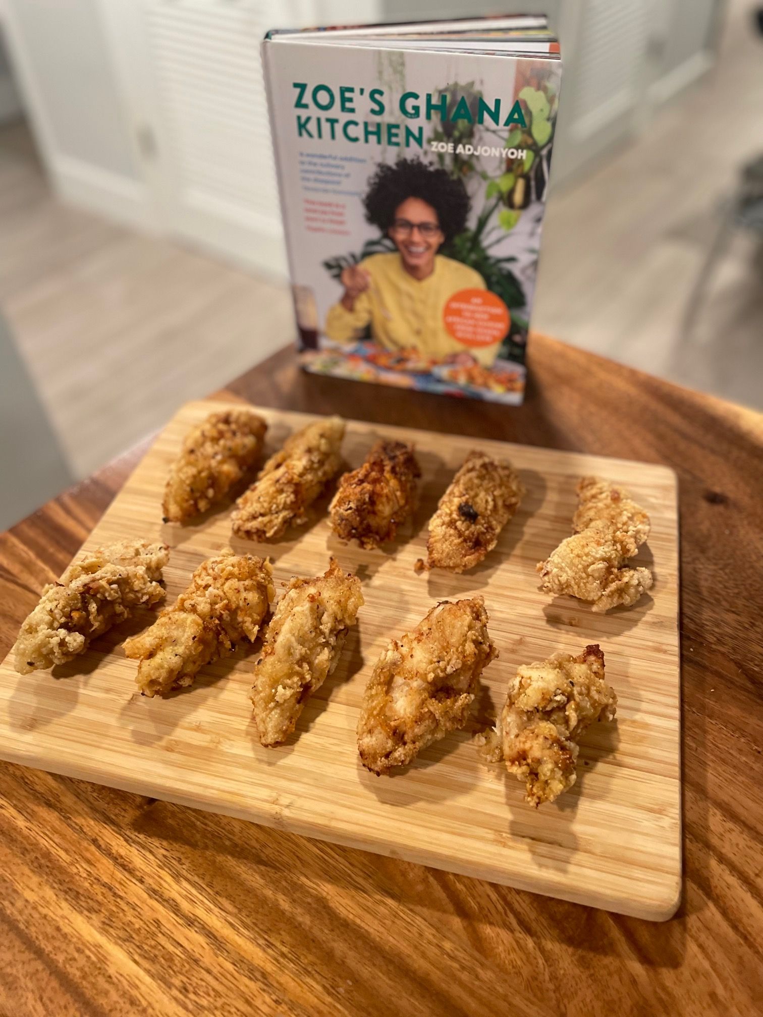 Fried chicken breast strips on a wooden cutting board in front of the cookbook Zoe's Ghana Kitchen