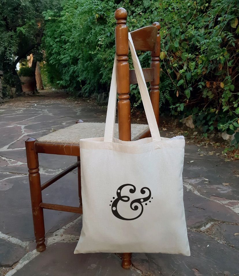 Image of a tote bag with an intricate ampersand design. The tote is hanging off a chair. 